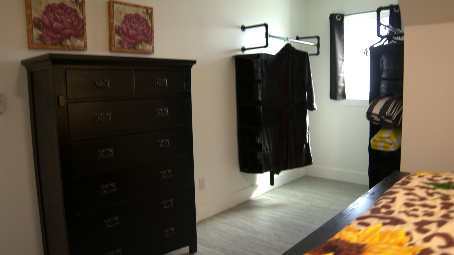 Bedroom dresser in dark wood under two rose paintings, with a clothes rail by the window